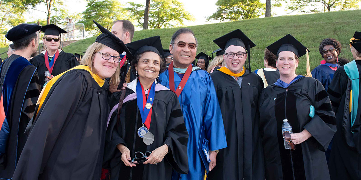 A group of faculty members dressed in academic regalia pose and smile outside at Commencement