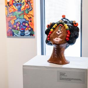 A clay bust sculpture of a person's head with stylized hair, colorful beads across the forehead, and red beads framing the face, displayed on a pedestal in an art gallery.
