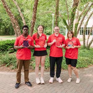 Four student ambassadors, wearing red shirts, hold up awards they have won.