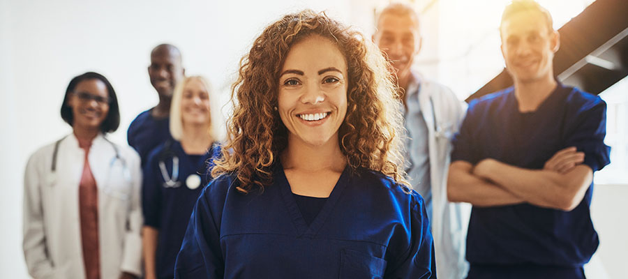 A health care worker in blue scrubs stands in front of a variety of other health care workers