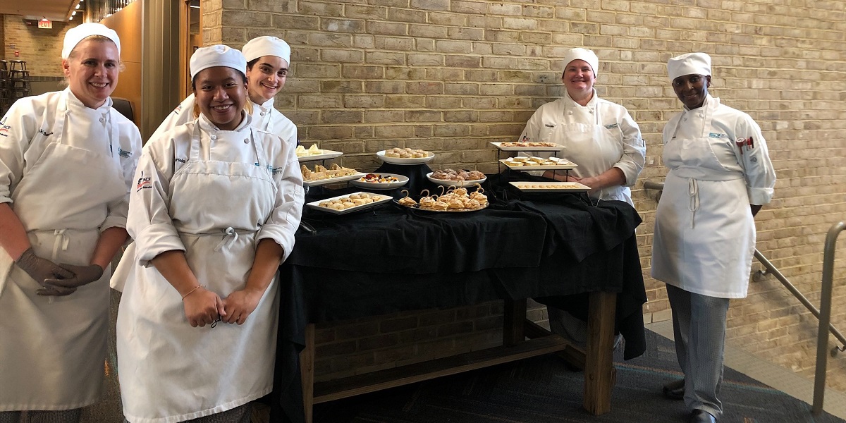 Five baking students stand next to a table of pastries.
