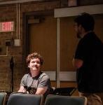 Two people conversing in a dimly lit auditorium. One young man sits behind a row of theater seats, looking up at a man standing in a black shirt.
