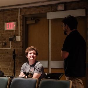 Two people conversing in a dimly lit auditorium. One young man sits behind a row of theater seats, looking up at a man standing in a black shirt.