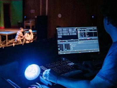A theater technician works at a lit control console in a dark booth while actors rehearse on a distant stage.
