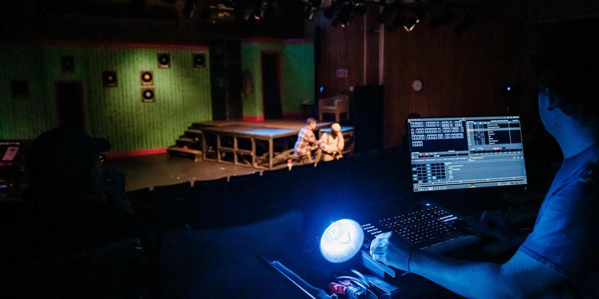 A theater technician works at a lit control console in a dark booth while actors rehearse on a distant stage.