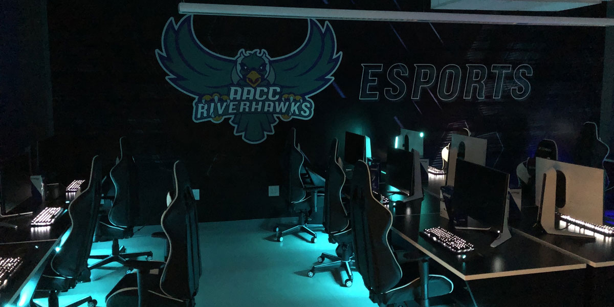 Image of AACC Riverhawks logo with Esports written on the wall. Image of gaming chairs with computers lined in a row.