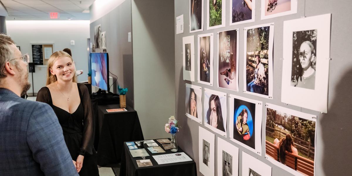 A student stands in front of their artwork on the wall, explaining it to another person.
