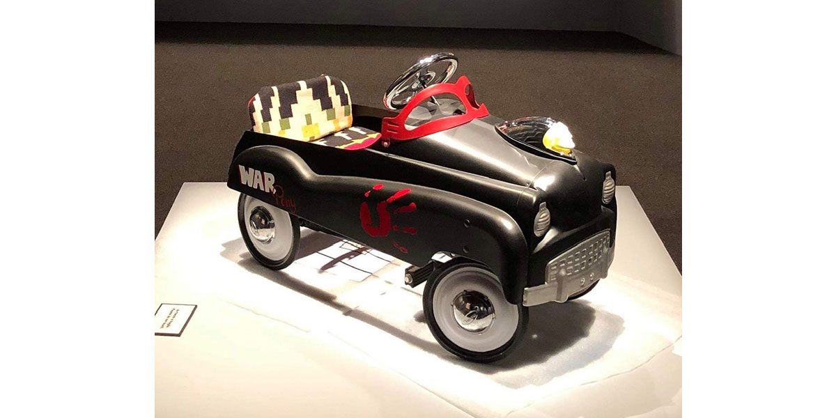 A black vintage-style toy pedal car sits on a museum pedestal. It is marked with 