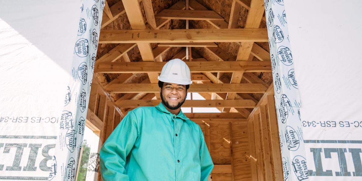 A student, wearing a hard hat, stands in the foundation of a building being constructed.