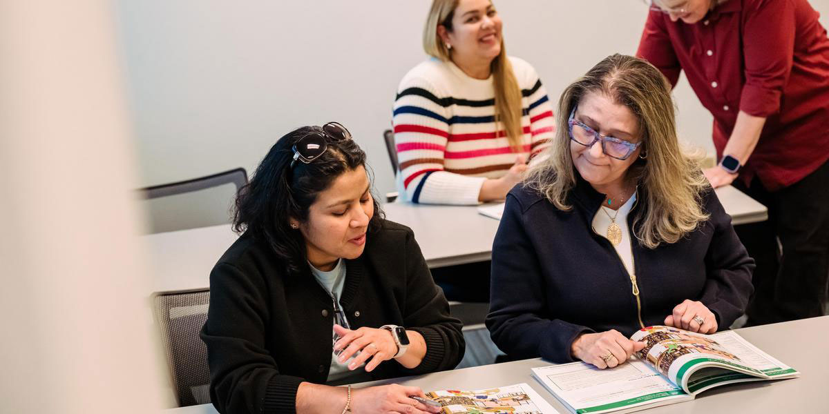 Two students seated next to each other, talking and looking at a textbook.
