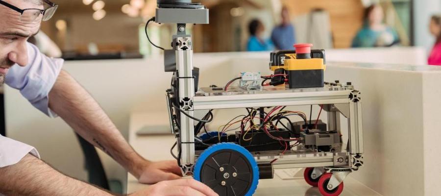 A man adjusts a wheeled robotics prototype with an exposed aluminum frame, blue wheels, visible wiring, and a red button.