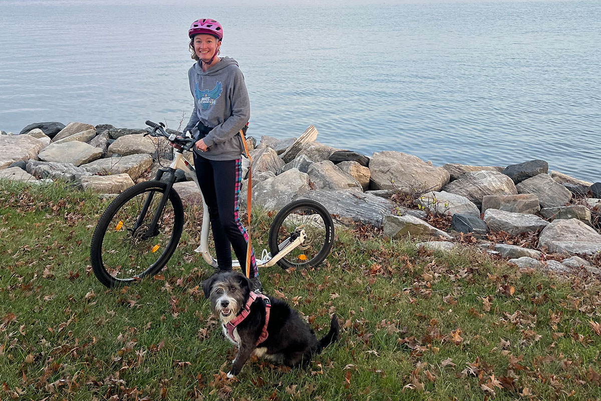 Candice Mayhill standing next to her kickbike with her dog. Water is in the background.