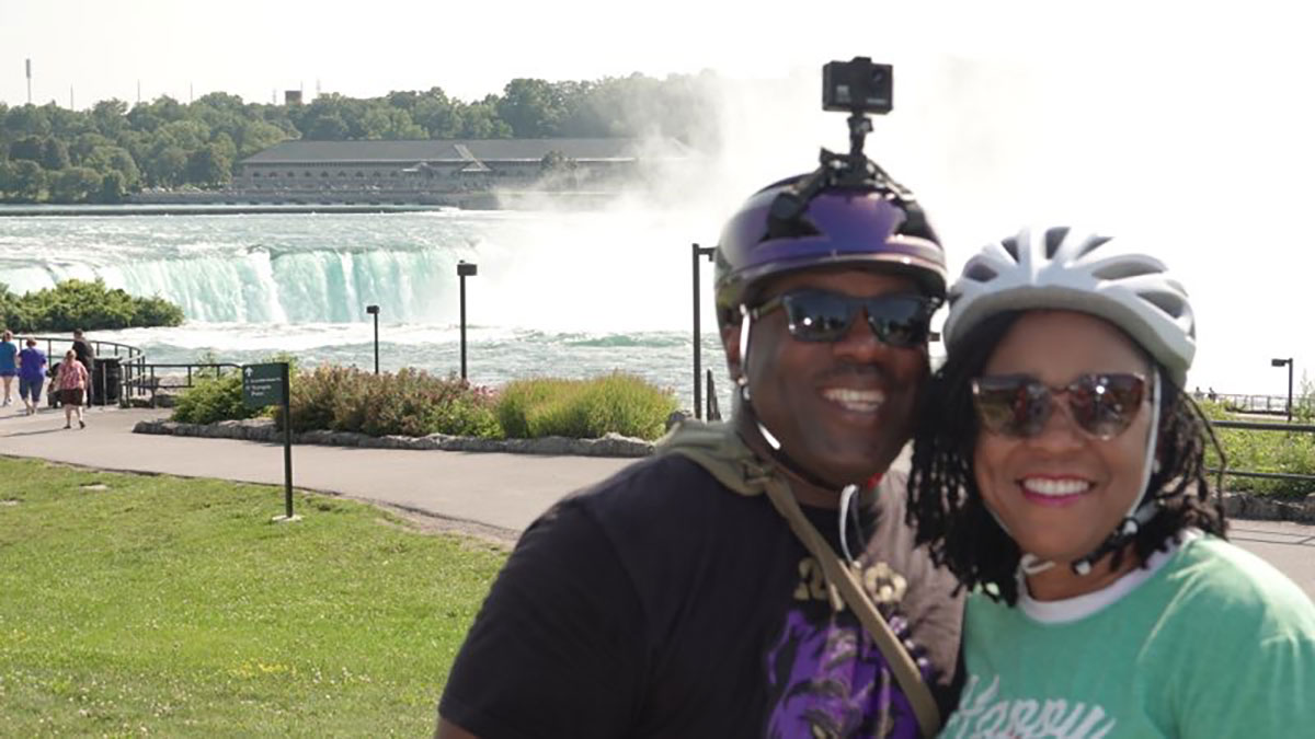 Tracey Short and partner standing in front of Niagara Falls. Both are wearing bike helmets.