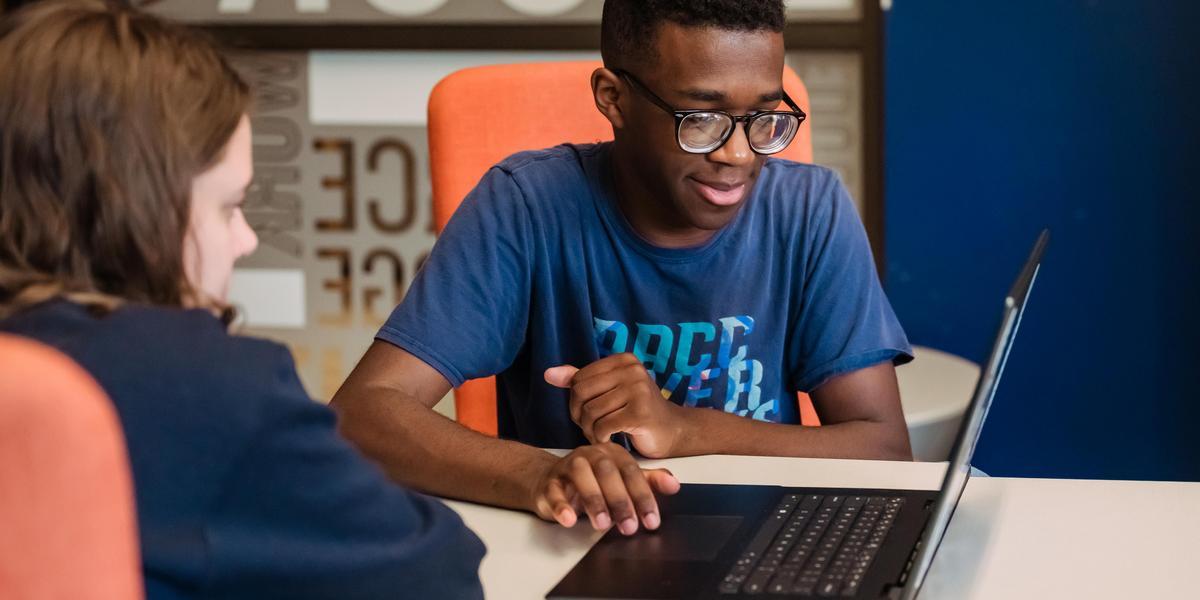 One student uses a laptop, showing the screen to another student.