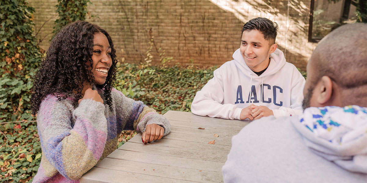 A group of students sits around an outdoor table on campus talking.