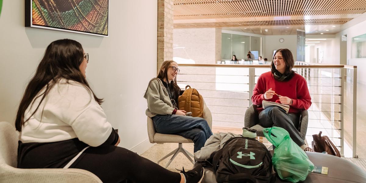 Three students sit in lounge chairs in a bright campus hallway, talking and smiling. Backpacks and a tote bag rest on the floor between them, and other students are visible in the background through a glass railing.