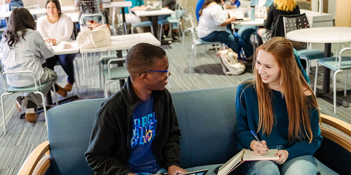 Two students sit on a couch and talk. They hold a laptop and a notebook. Other groups of students sit at tables behind them.