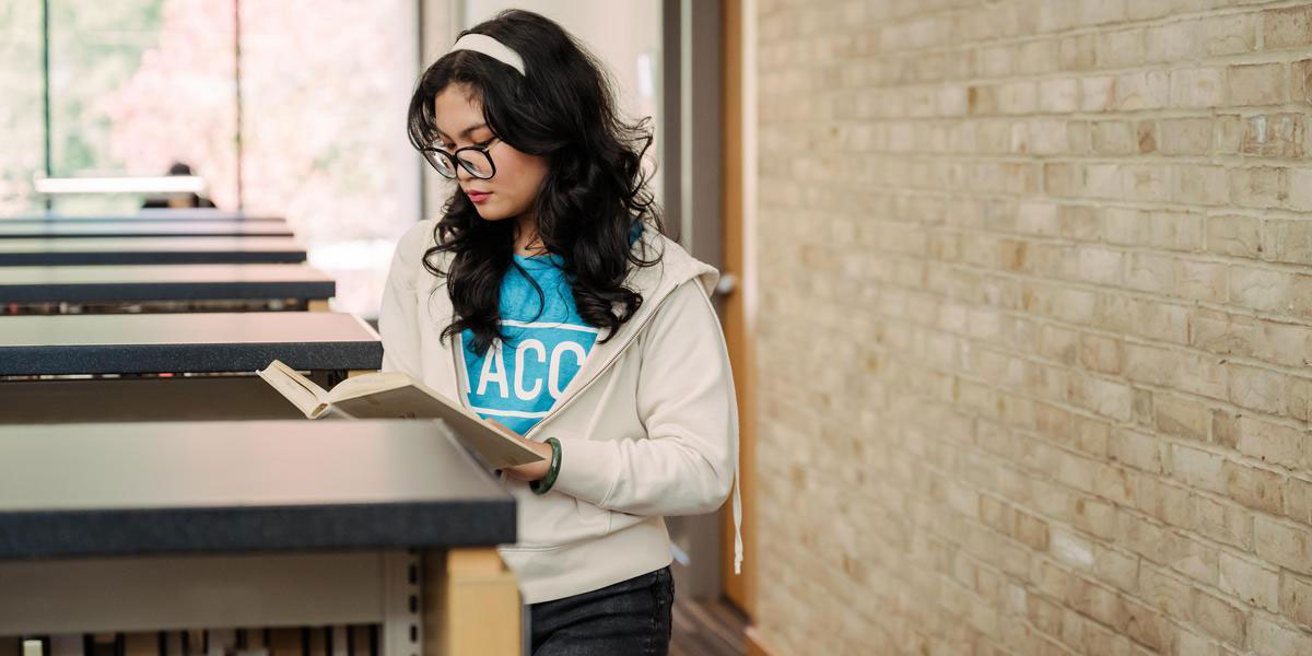 A student in an AACC T-shirt reads a book while standing near library shelves.